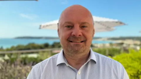 Kristjan Nommik is wearing a striped shirt and had a beard. St Aubin's bay is in the background of the photo