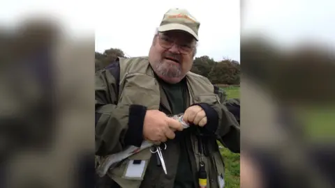Alan Graham pictured holding a fish. He is wearing a khaki hat and fisherman's jacket. He has a grey beard and wears circular silver framed glasses