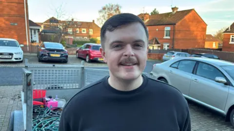 A brown haired man with a moustache and a black jumper