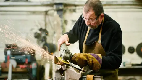 Brian Fell/Adrian Lambert Brian Fell (L) has short cropped brown hair, brown glasses and wears a black sweat shirt and stained khaki apron and is using a grinder which sparks are flying off.