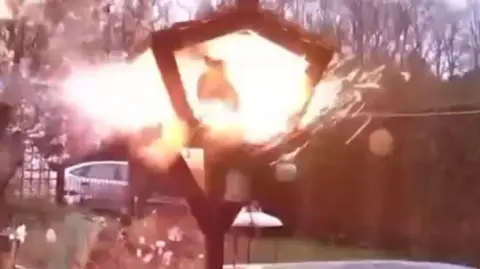 James Garnor A triangular wooden bird table is engulfed by an orange flash. Sparks appear to be flying off the bird table. A cat is seen surrounded by the orange flash.