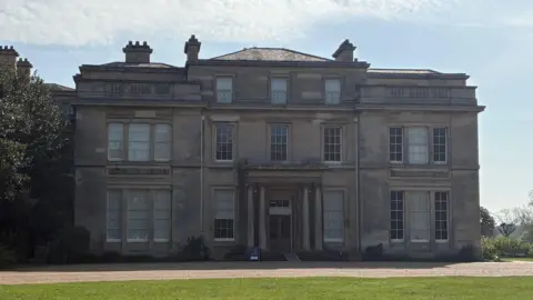 Kevin Shoesmith/BBC A three-storey, stone-built mansion with a lush lawn in front it it. The entrance contains four pillars.