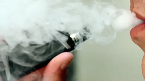 A man is using a black vape. There is smoke coming from his mouth.