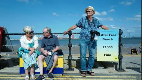 Lee Shulman An older man wearing sunglasses, a summer shirt, hat and jeans and sandals, leans on a black fence on a promenade on a sunny day, next to an older couple. A camera is hanging around his neck 