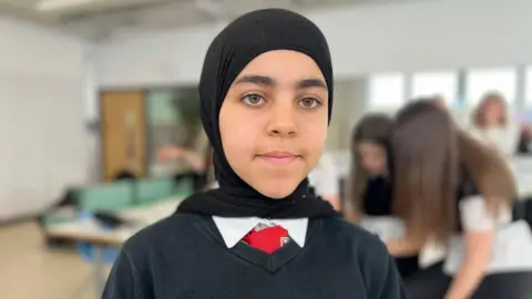 Razan is looking at the camera. She wears a school uniform of a black jumper, white shirt and red tie. She has a black headscarf over her hair.