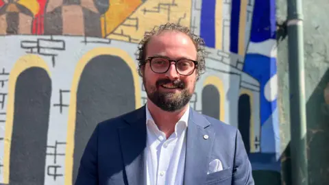 Sue Dougan/BBC Head and shoulders image of Ross Martin standing in front of a colourful wall. He has short curly black hair, a beard and is wearing dark rounded glasses. He is wearing a navy suit and light shirt.