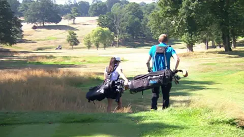 Golfers walk away from the camera down a hill on a sunny day at Stoneham Golf Club. The landscape features trees and areas of long grass.