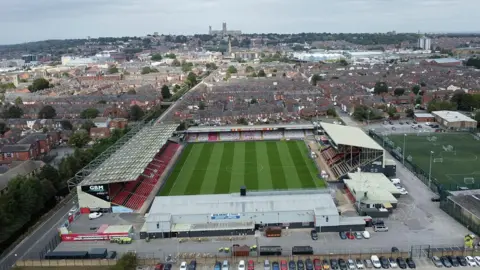 A drone view of a football stadium with four stands of various sizes and a green pitch. Behind it are rows of terraced houses and looming on a hill in the background is a large cathedral.