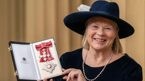Getty Images Dame Ann Limb, who wears a navy broad-rimmed hat with a white feather, has a blonde bob and wears a navy long-sleeved top with a pearl necklace, holds up her medal and smiles in a photo 