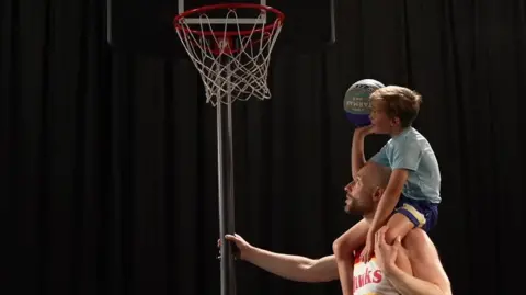 Six-year-old Luther on his dad's shoulders aiming to shoot the basketball through the hoop