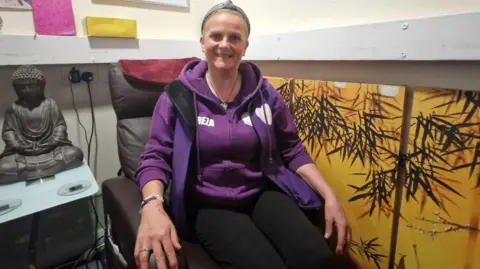 LDRS A woman wearing a purple hooded, zipped-up jumper sits in a brown leather chair, on the left side is a statue of Buddha and on the right side a series of pictures of branches with a yellow background hangs on the wall