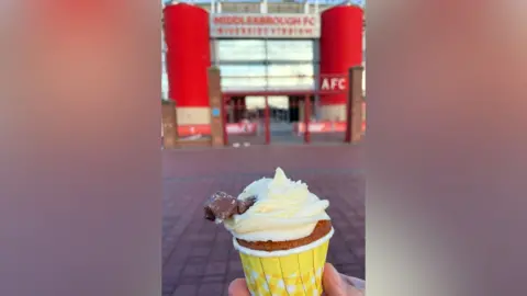 Karen Campbell A cupcake in a yellow paper case with white icing and a small chocolate flake sticking out the top, held in front of the entrance to Middlesbrough FC's Riverside Stadium, which is flanked by two large, red pillars.