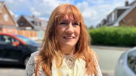 Maureen O'Bern has long ginger hair and is smiling at the camera. She is wearing a white lace top and yellow scarf. 