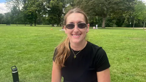 Daisy Robson sits in a green field staring at the camera with sunglasses on and wearing a black T-shirt.