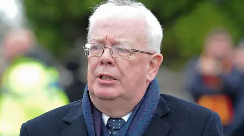 A man with grey hair and glasses is wearing a blue coat, blue scarf, white shirt and blue and white spotted tie