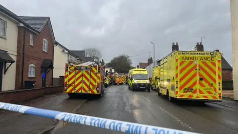 A fire engine and three ambulances are parked on a residential road while police tape prevents people from entering the scene. 