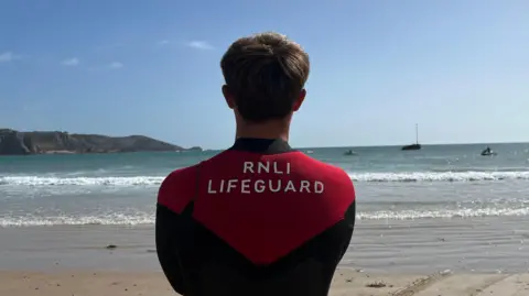 A lifeguard in a red-and-black RNLI wetsuit stands at St Brelade's beach facing the sea. Gentle waves roll in under a clear blue sky, and a couple of lifeguards on skis are visible offshore. Low cliffs stretch along the coastline in the distance.