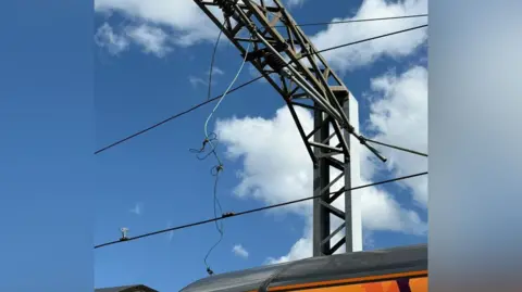 Network Rail Loose cables hanging near a steel beam with the roof of a train showing below them. The sky is blue overhead with some white clouds.