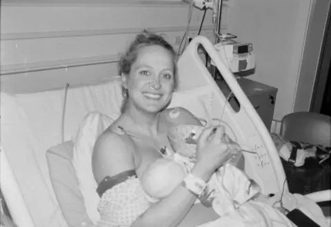 Francis Sanders Naomi holding Ester after her birth. Naomi is sitting up in a hospital bed with two wires attached to her shoulders and is smiling at the camera wither her hair tied back.