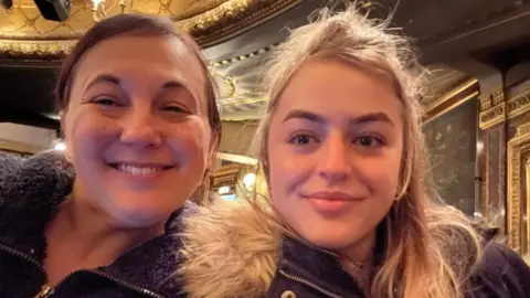 MARIA SOLECKI Maya is on the right and her mum Maria is on the left. Maya has blonde hair, Maria has dark hair. Both of them are in what seems to be a theatre auditorium.