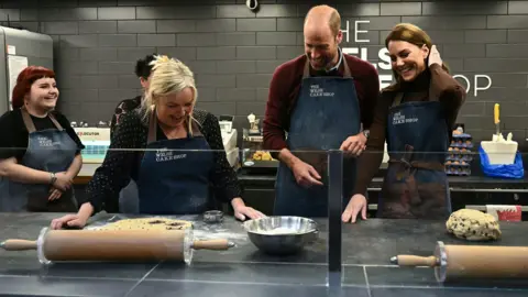 Ben Stansall/PA Wire The Prince and Princess of Wales stand with denim aprons that say The Welsh Cake Shop on in white lettering. They are laughing as the owner of the shop, Theresa Conner, shows them how to make Welsh cakes. On the counter before them is dough, rolling pins on both sides, and a silver bowl with flour in. 