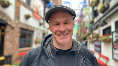 A man smiles into the camera. He is wearing a dark cap, dark coat and dark shirt. He is standing in the street with the iconic umbrellas behind him.