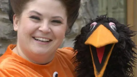 S4C Rebecca Harries has brown hair which is tied back. She is wearing an oragne costume and holding a black bird puppet. She is smiling and looking at the camera.
