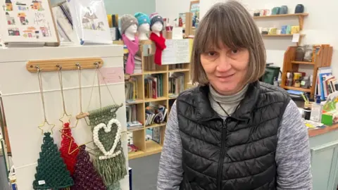 BBC Helen Fallows has short brown hair and is standing in her shop in front of a number of items, like Christmas decorations. She is wearing a black gilet and grey top. 