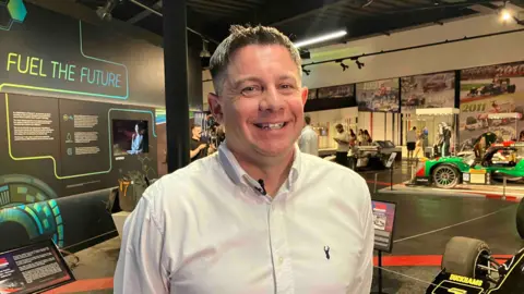 A man in a pale blue shirt stands in a room with exhibitions of cars smiling at the camera.
