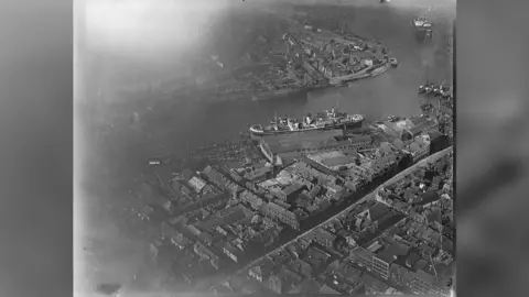 Historic England A black-and-white aerial shot of a ship and masses of houses along a large winding river.