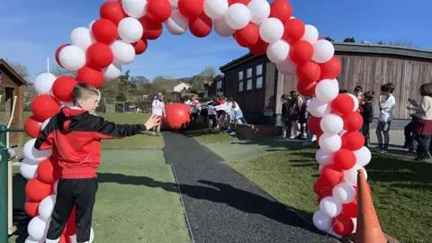 A red and white balloon arch in front of a demountable classroom on the school field. A crowd of children is lining the route.