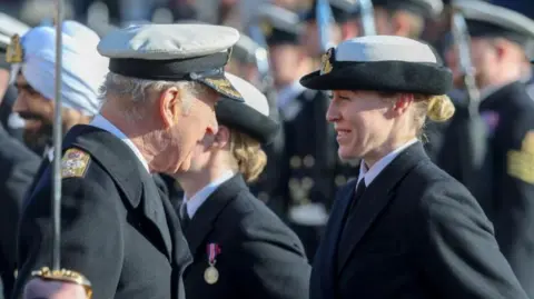 Chris Jackson/Getty The king talks to a young woman whose blonde hair is in a bun under her hat. She has a navy uniform and tie and is smiling.