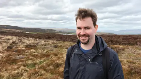 Gwyndaf Hughes/BBC A man wearing a navy blue coat standing on a hillside with brown heathland behind him