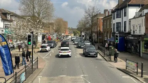 Nigel Thompson/Geograph A general view of Epping High Street in the town centre. Cars are being driven either side of the road, which is lined by shops and trees. This picture has been taken from a tall vantage point and shows shoppers by a traffic light crossing.