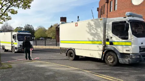 Two large white police trucks parked in front of a red brick building. A police man, dressed all in black, is stood between the trucks, back to camera, holding white and red cordon tape in a residential street. There are trees and railings visible in the background.