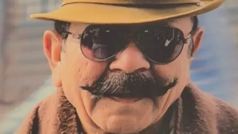 A man with a black moustache wearing a yellow hat and sunglasses. The background behind him is blurred.