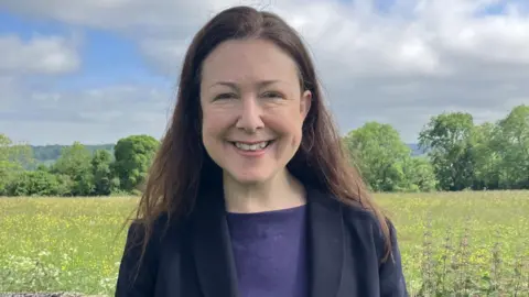 Green Party Chloe Turner standing in a green field boardered with trees. She has long brown hair and is wearing a dark top and suit jacket and smiling at the camera.