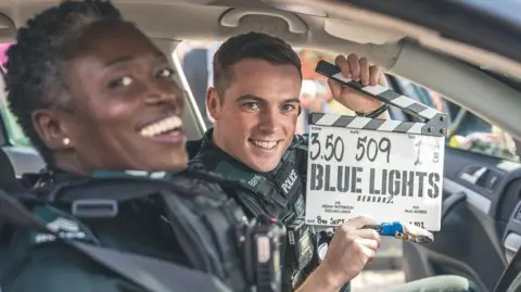 Two cast members of the series Blue Lights, one woman and one man in PSNI uniforms in a police car. The man is holding a Blue Lights clapper board, both are smiling     