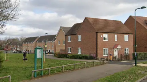 Google A row of red brick two and three houses in Orton Northgate. In front of them is stretch of grass with a dog walker on it. 