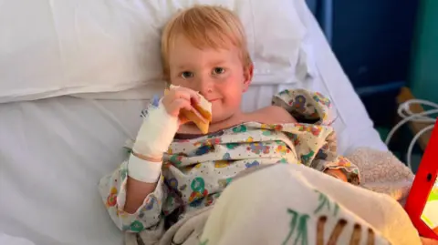 Logan is sat in a hospital bed with a sandwich and a blanket. 