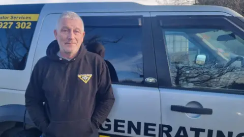 Steve Batey Steve Batey is standing with his back to the side of a grey Land Rover. You can see the word generating written on the vehicle. He is in his 50s with grey hair and wearing a blue top with a yellow company logo.