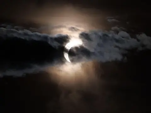 BBC Weather Watchers/Bram The partial eclipse captured in Marske-by-the-Sea. The sky is dark and there are clouds across the partially covered sun.