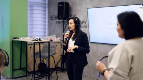 Nataliia Zaslavska Sonia Kumar is giving a speech whilst holding a microphone and wearing headphones for translation. She is wearing a black suit with white blouse. Behind her a large screen is mounted on a grey wall with the word innovation and physiotherapy written in relief.