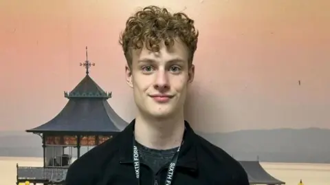 Jack, wearing a black top and a sixth form lanyard. He has curly brown hair. He is stood in front of a picture of Clevedon Pier.