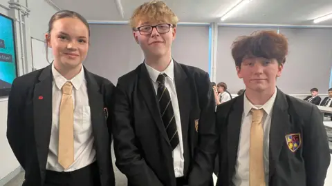 Three teenagers in black school uniforms with white shirts and yellow and black ties stood in classroom