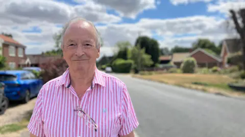 Owen Sennitt/BBC A man wearing a pink and white-striped shirt, with glasses being held through a button slot, looks towards the camera while stood next to a road, which is out of focus