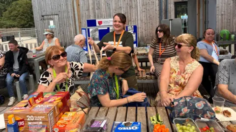 Right At Home Central Wiltshire A group of people sitting on wooden and metal picnic benches outside. There is an array of picnic food on the table. Some people are wearing orange lanyards.