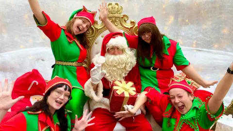 A Santa Claus on an ornate chair with a white beard holding a parcel with yellow ribbon. He is surrounded by women in green and red and they are all smiling.