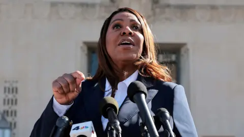 A woman wearing a suit speaks in front of microphones.