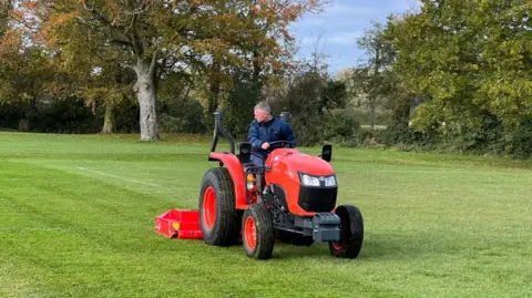 A man cuts the grass of a football pitch on a red tractor with trees in the background.